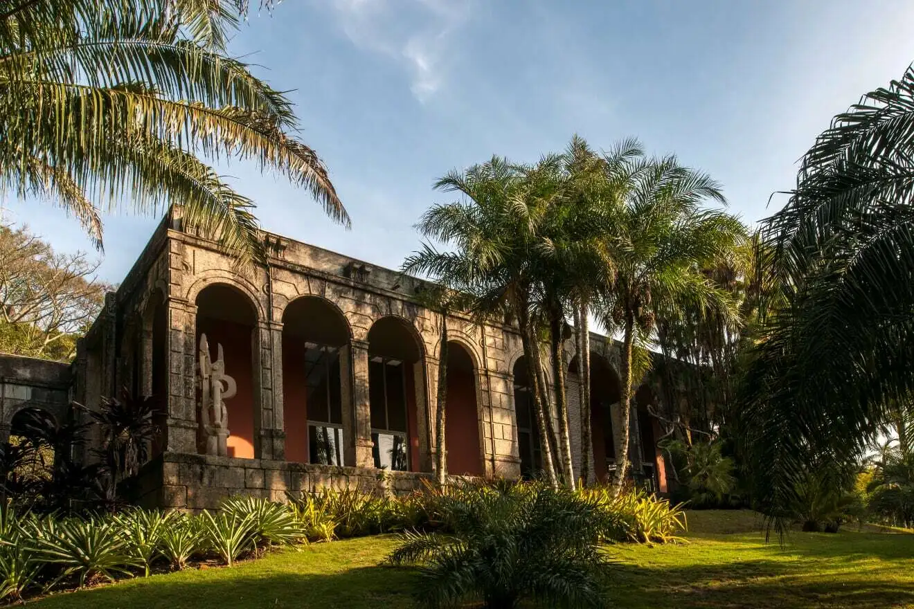 Photo d'une partie du jardin de Sitio Roberto Burle Marx. On y voit un bâtiment avec une architecture ancienne divers massifs de plantes exotiques tels que des palmiers. On peut également voir une structure surement par l'architecte paysagiste Roberto Burle Marx